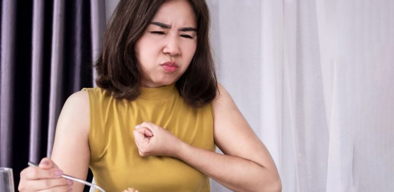 A woman in a tan colored shirt, holding a fist to her chest. She is having trouble swallowing the food she's eating.